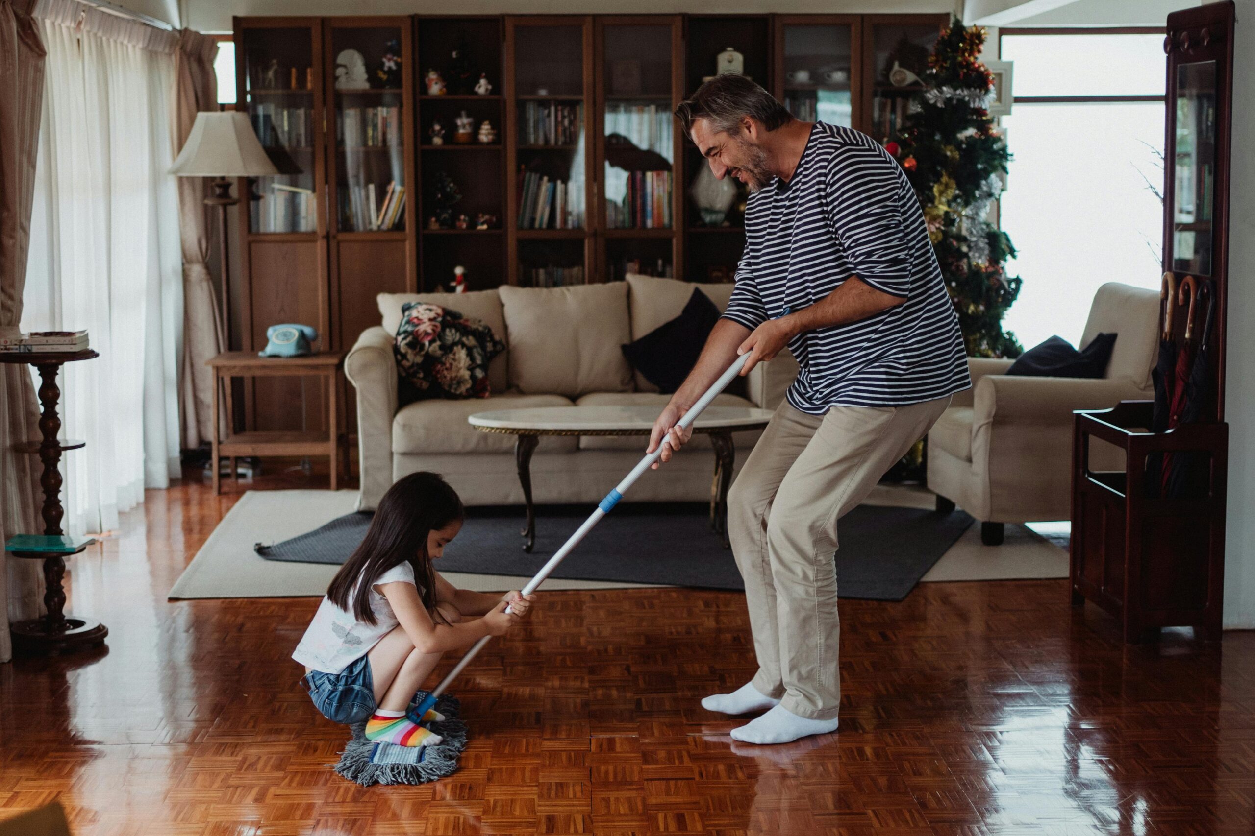 A playful family moment as a father and daughter mop the living room floor together, enjoying quality time.